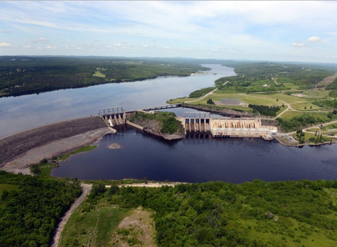 Aerial view of the Mactaquac Generating Station looking towards the arm (North-Northwest). The left side of the dam is earthen, the sluiceway central, and the spillway, turbines, and powerhouse on the right. Mactaquac Road is seen on the top of the embankment and crosses the rest of the river upstream of the turbines. 